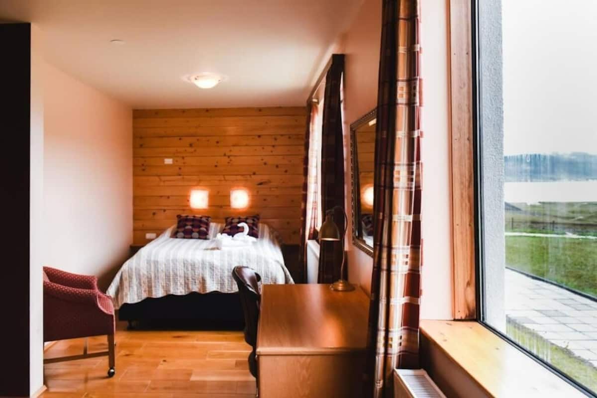 Un dormitorio con piso de madera, una cama con cubrecama blanco, una silla roja, un escritorio de madera y un ventanal con vista a una masa de agua.