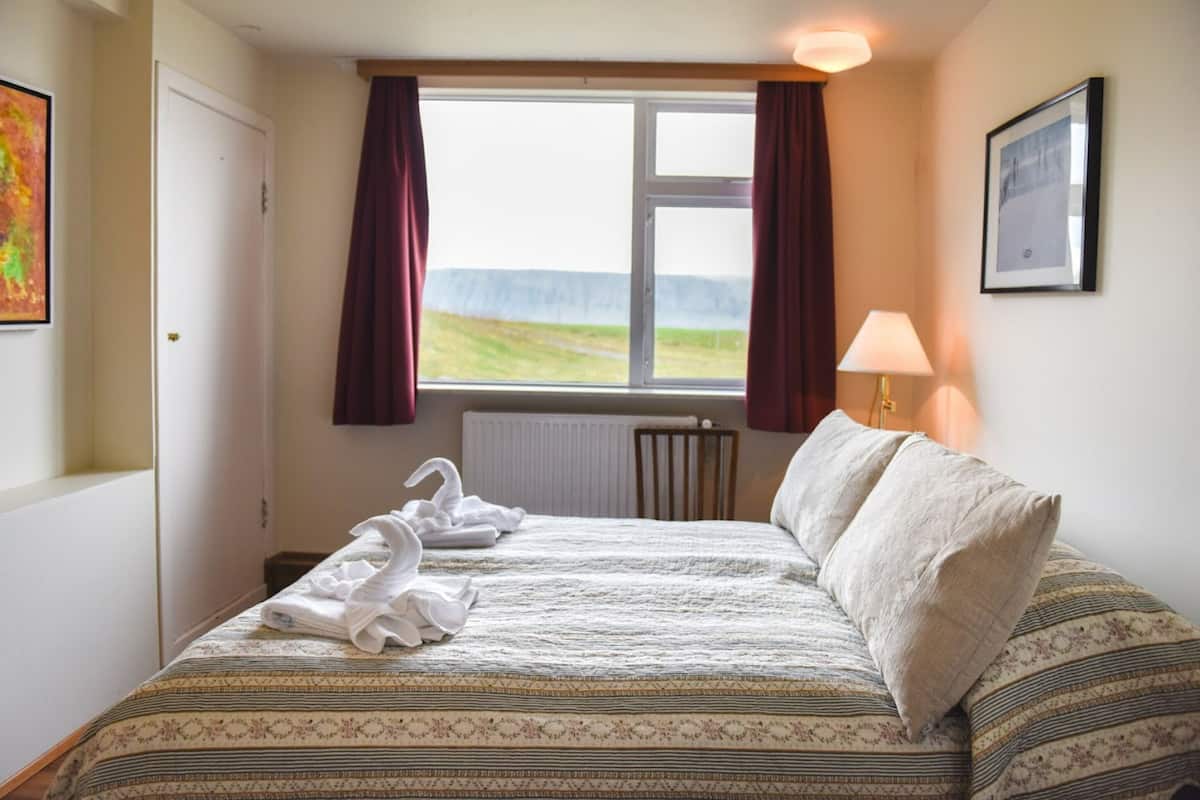 Una habitación de hotel con cama, ventana con cortinas, radiador, lámpara y un cuadro en la pared.