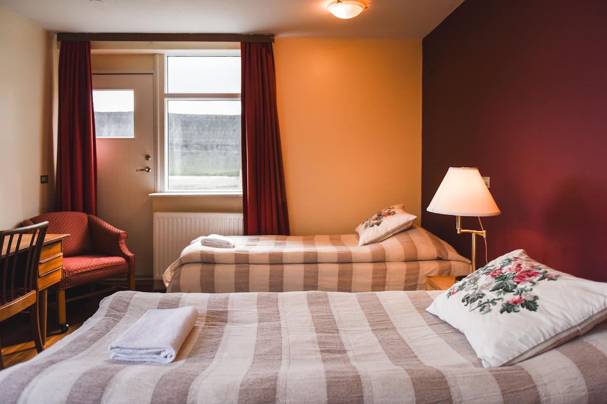Habitación de hotel con dos camas, un escritorio, una silla, un ventanal con cortinas y una lámpara.