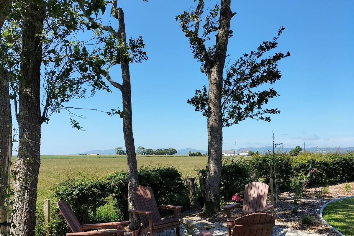 A seating area with wooden chairs and a table, surrounded by trees and a gravel path, overlooking a green field and distant mountains.