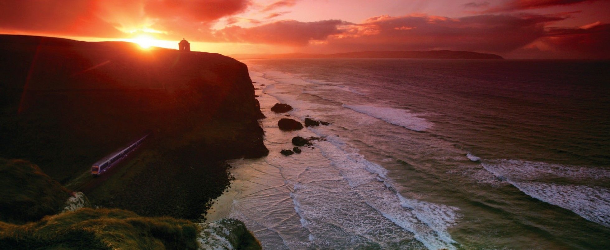 A coastal sunset with a lighthouse on a cliff, waves crashing, and a train track.