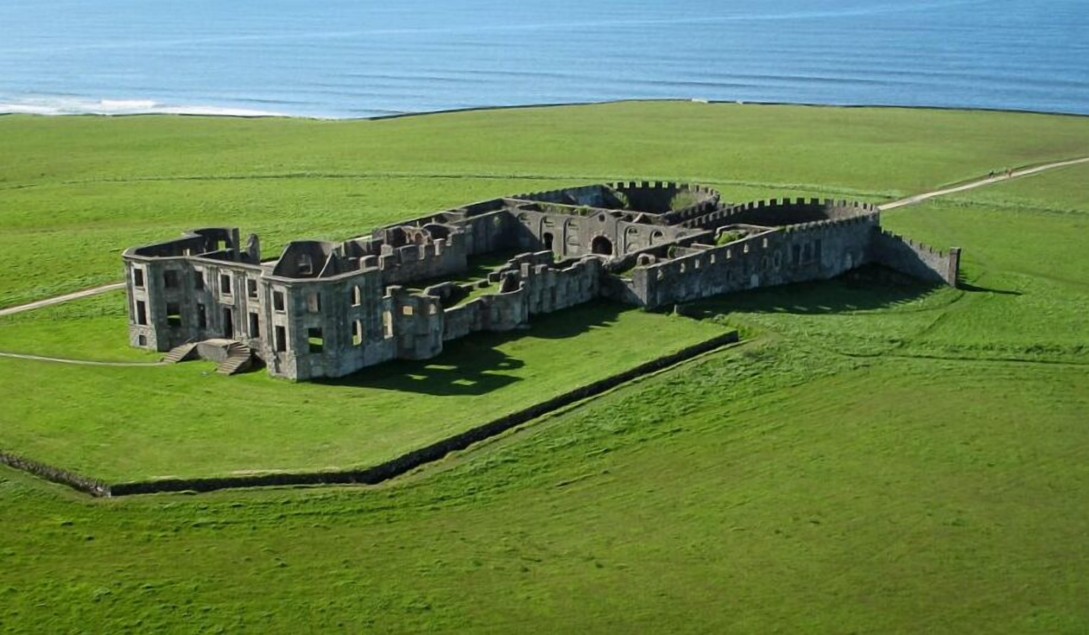 Vista aérea de un castillo histórico de piedra con múltiples torres y un foso, rodeado de campos verdes exuberantes y una masa de agua al fondo.