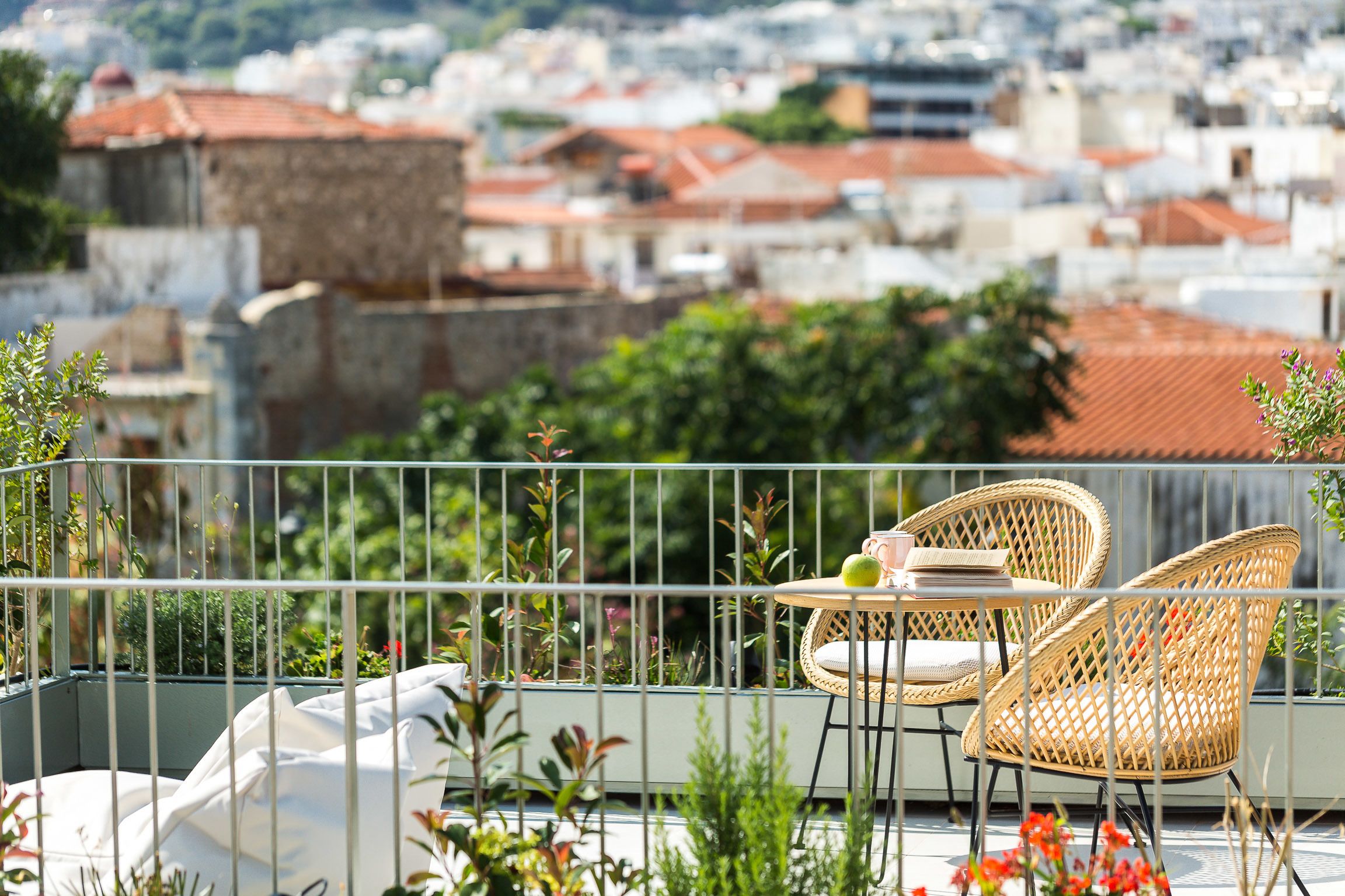 A balcony with wicker chairs and a small table, overlooking a cityscape with red-roofed buildings.