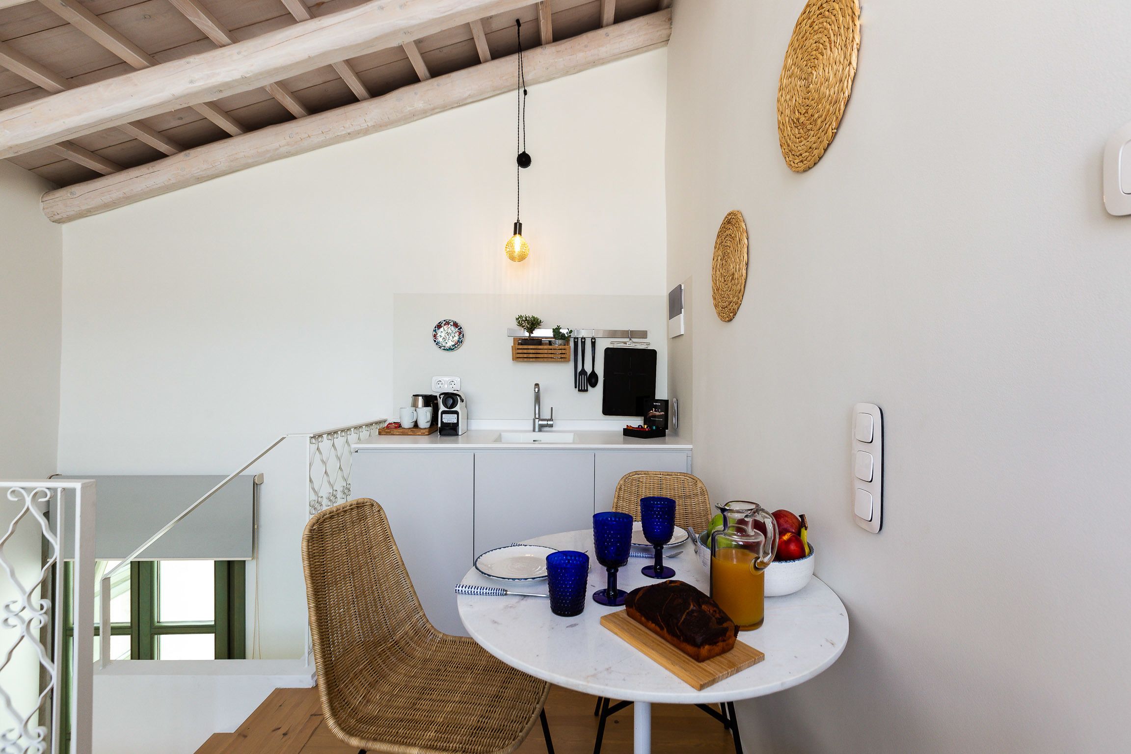 A dining area with a round table, wicker chairs, and a wooden bench. A kitchen with white cabinets and a hanging light fixture is visible in the background.