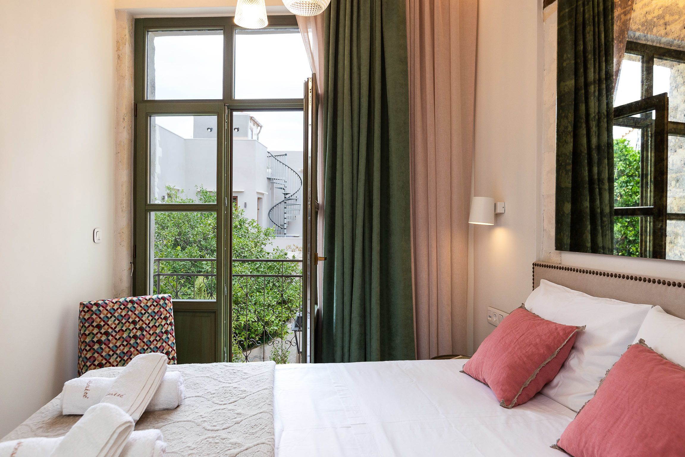 A hotel room with a bed, a window with curtains, a view of greenery outside, and a patterned pillow.