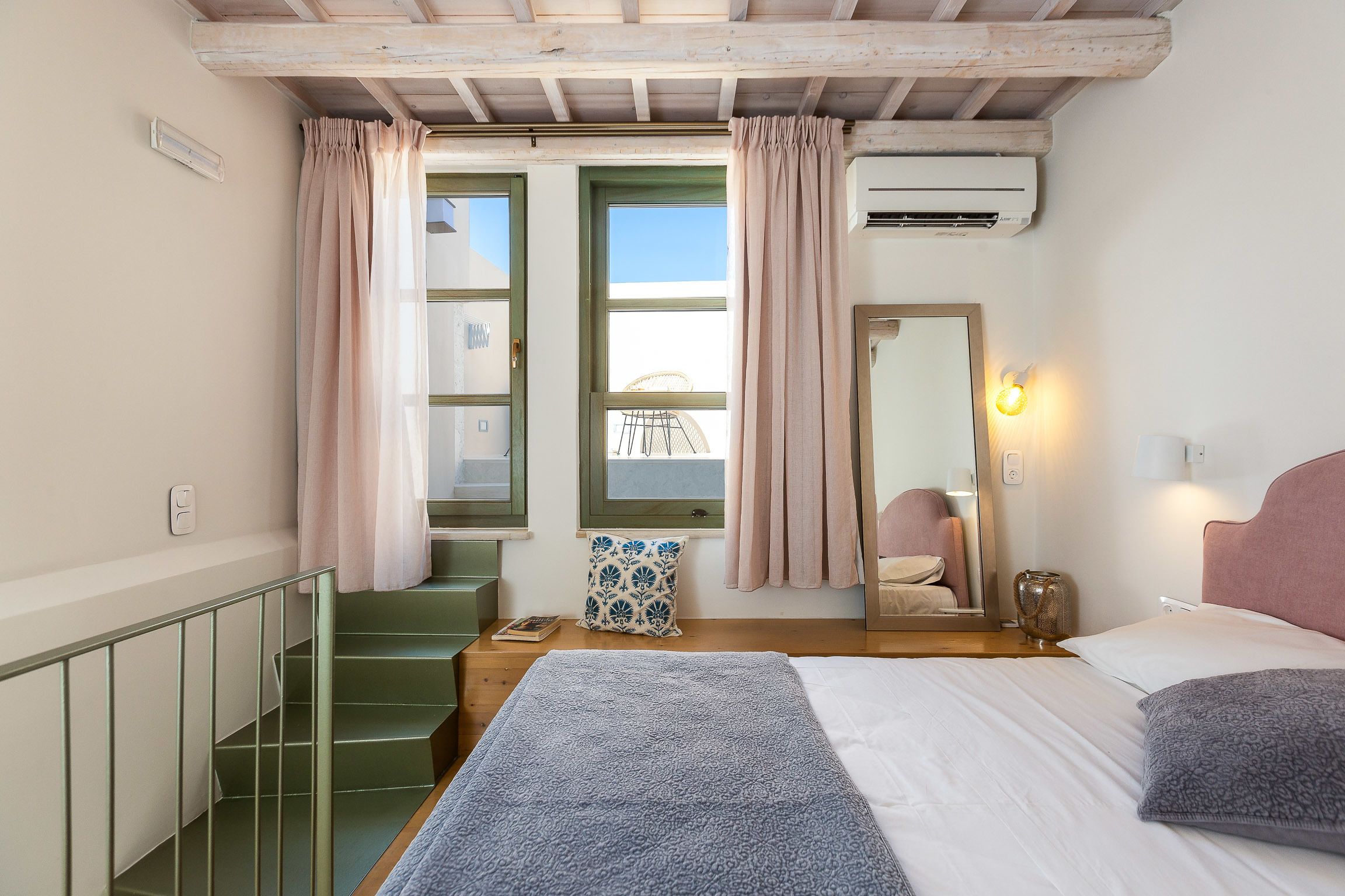 A bedroom with a bed, a nightstand, a window with curtains, a mirror, and a staircase.