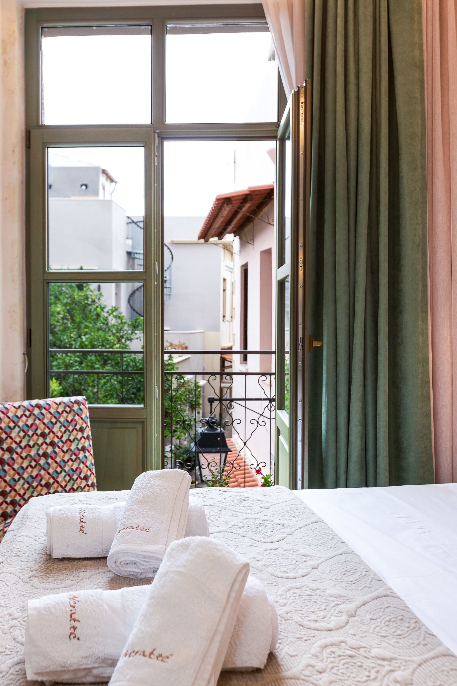 A hotel room with a bed, a window with curtains, and a view of buildings and greenery outside.