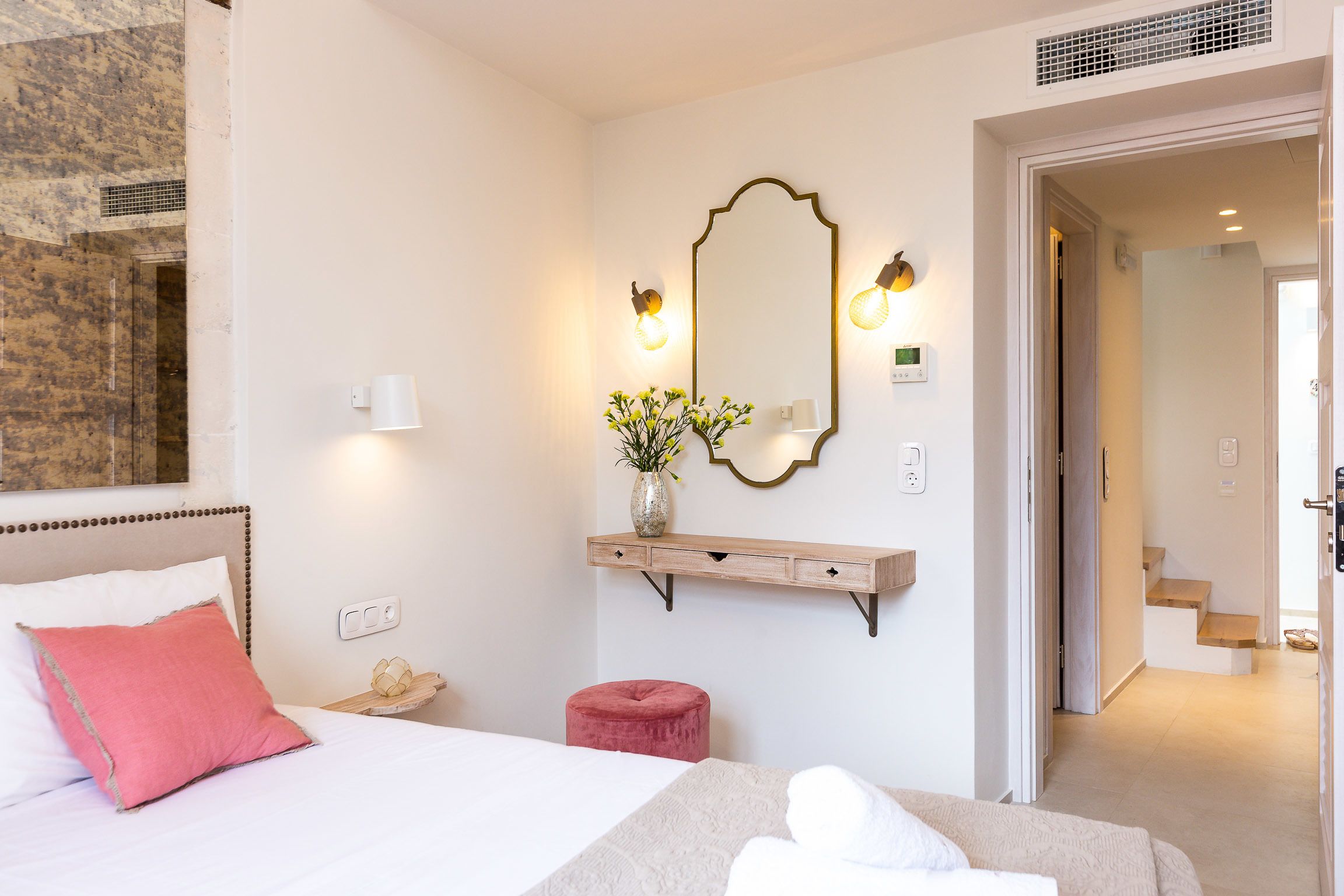 Una habitación de hotel con una cama, un cojín rojo, un pequeño taburete rojo, un espejo con apliques de pared y una estantería de madera con un jarrón de flores.