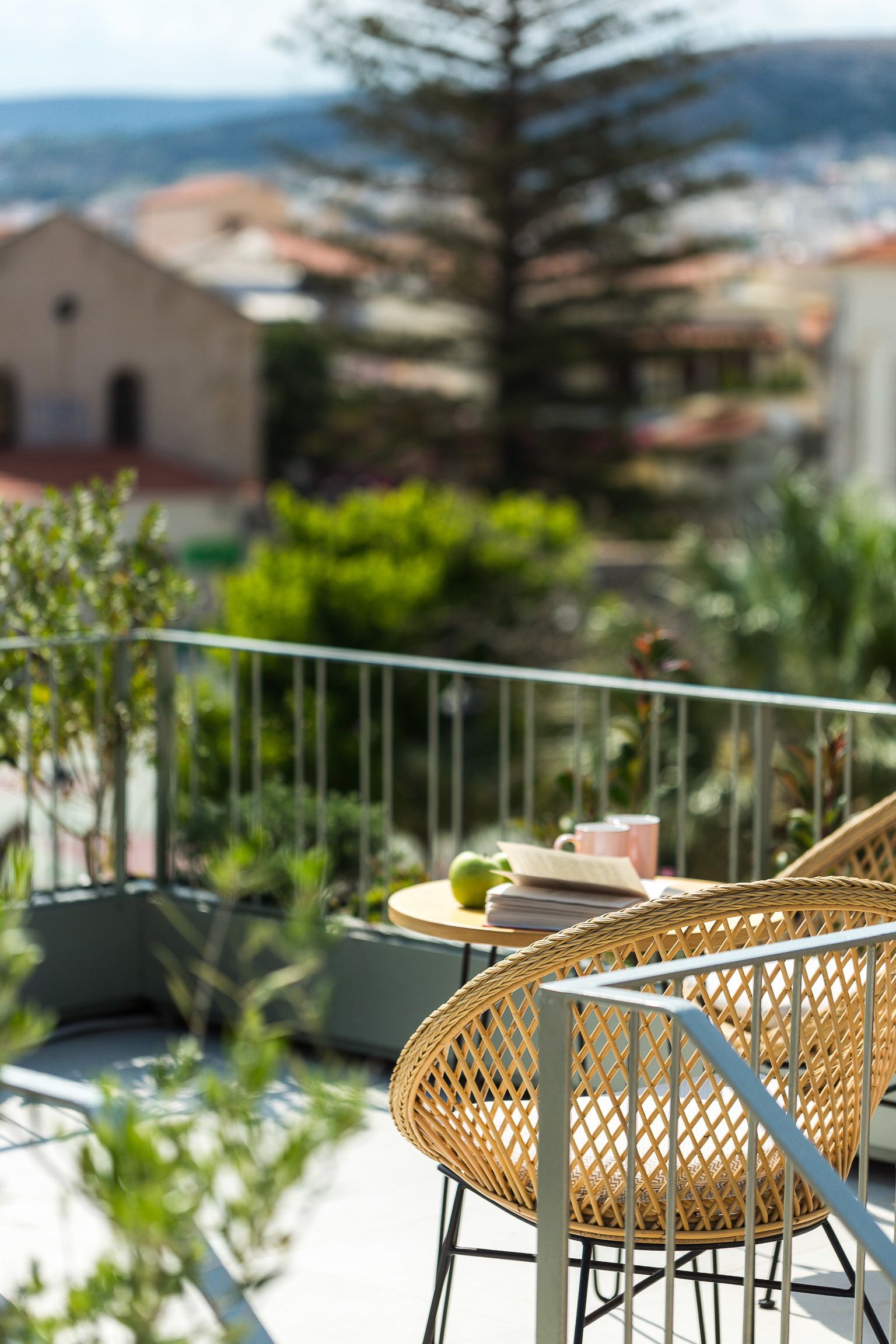 Un balcon avec des fauteuils en osier, une petite table avec un livre et une tasse, et une vue sur les bâtiments et les arbres en arrière-plan.