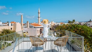 A balcony with wicker chairs and a table, overlooking a cityscape with buildings and a tall tower.