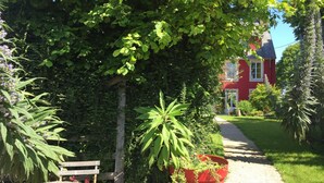 A red house with a garden path leading to it, surrounded by greenery and a wooden bench. - L'Ermitage (Saint-Vaast-la-Hougue)