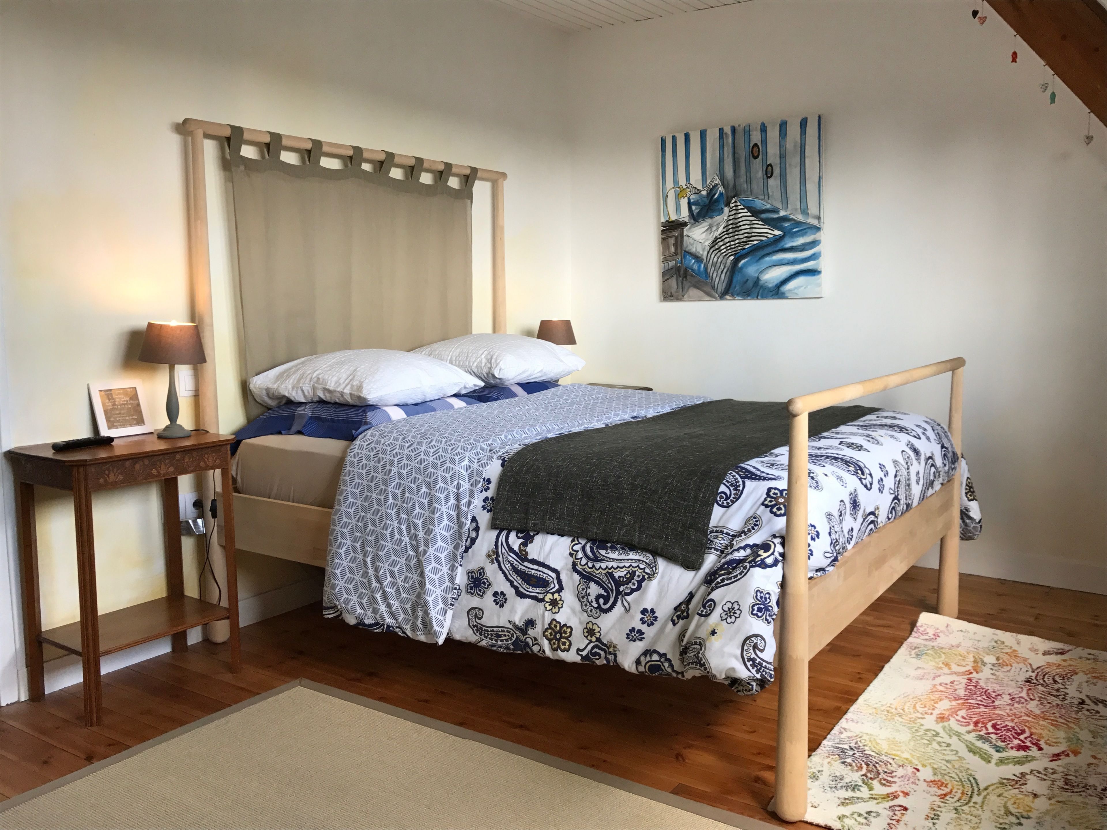A bedroom with a wooden bed, a nightstand with a lamp, a framed picture, and a painting on the wall.