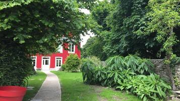 Ein rotes Haus mit weißer Tür und Fenstern, umgeben von üppigem Grün und einer Steinmauer.