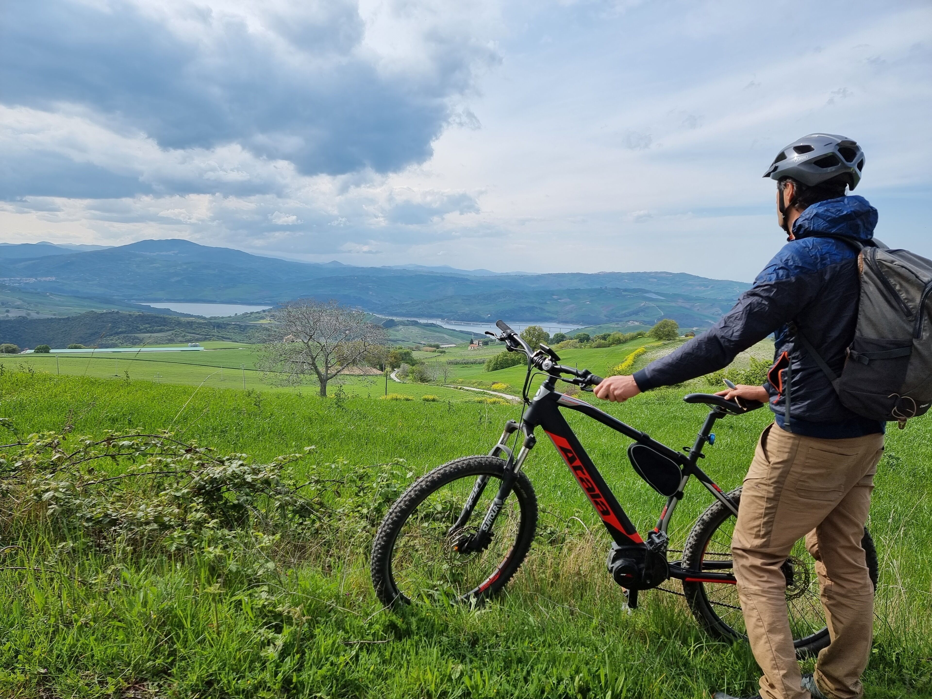 A person standing next to a mountain bike on a grassy hill with a view of distant mountains and a lake.