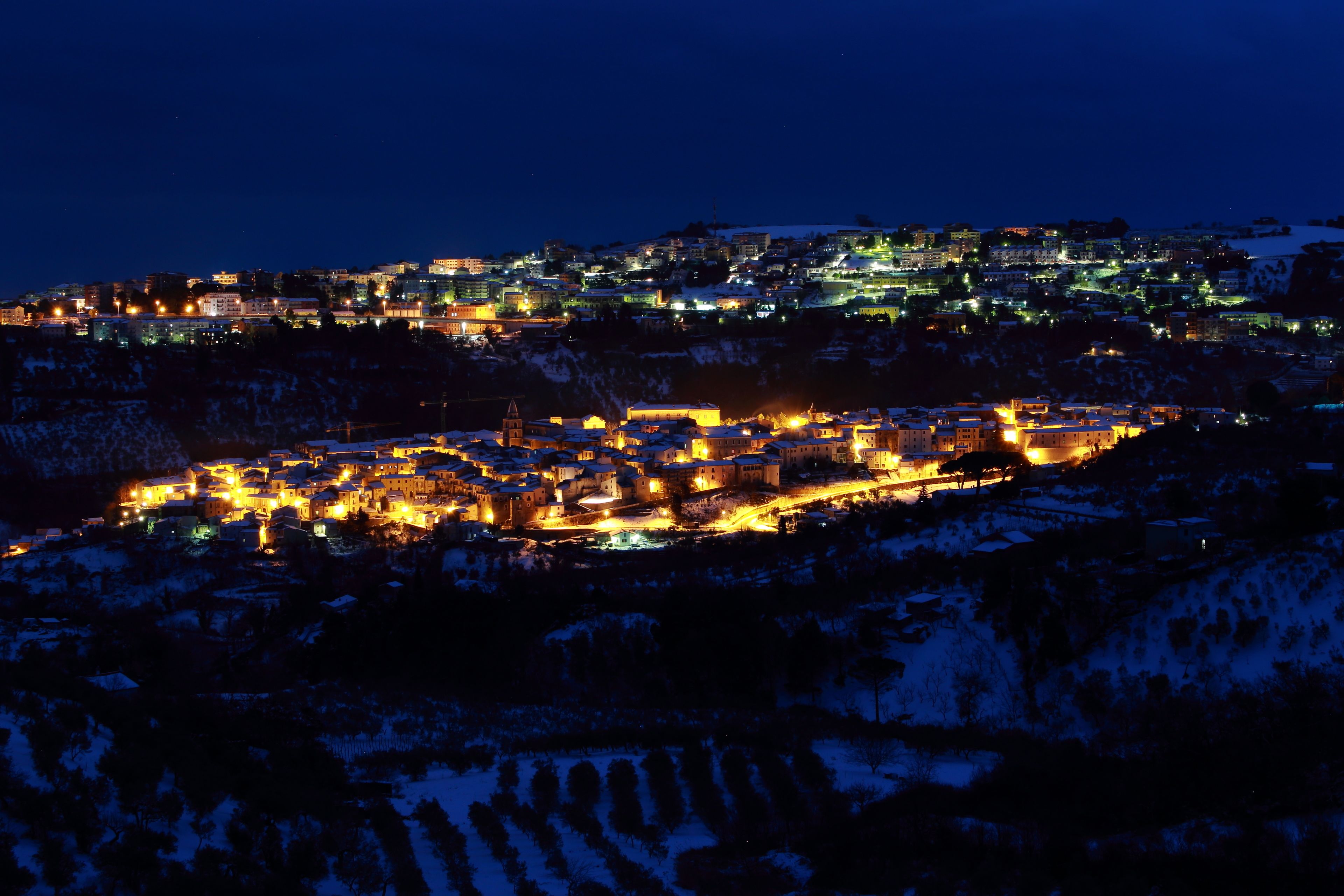Un pueblo cubierto de nieve por la noche, con edificios iluminados y una imponente aguja de iglesia.