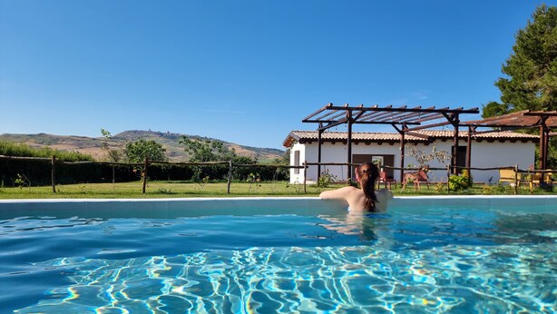 A clear pool with a person swimming, surrounded by a house with a pergola and a view of hills. - Agriturimo Essentia Dimora Rurale (Larino)