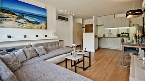 A modern living room with a large sofa, a small table with a vase, and a kitchen area with white cabinets and a dining table. - BlueApart Nadmorski Park Jastarnia (Jastarnia)