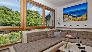 A modern living room with a large window, a sofa, a small table, and a painting of a beach scene. - BlueApart Nadmorski Park Jastarnia (Jastarnia)