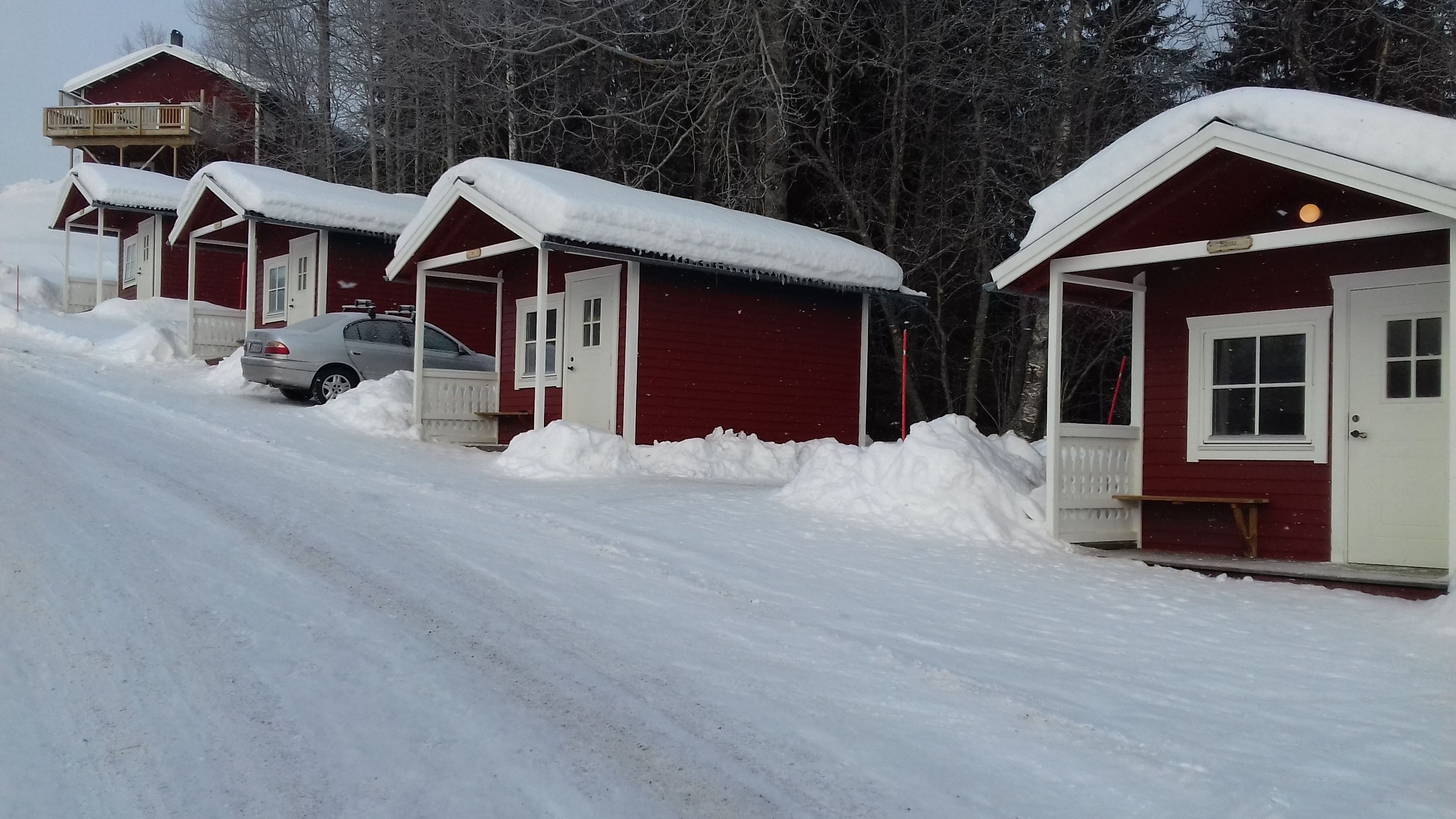 A row of red cabins with snow-covered roofs and a car parked in front.