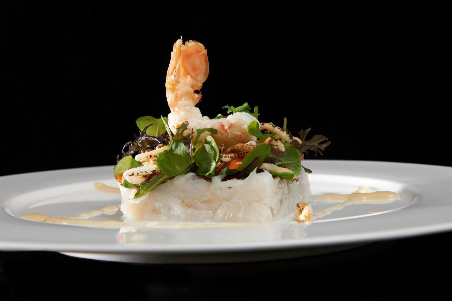 Une assiette blanche contenant un plat de fruits de mer, notamment une crevette, des légumes verts et, éventuellement, quelques champignons, le tout sur un fond sombre.