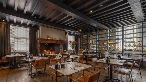 Un restaurant doté de planchers en bois, de poutres apparentes et d’une cheminée en briques. Les tables sont dressées avec des verres à vin et des couverts.
