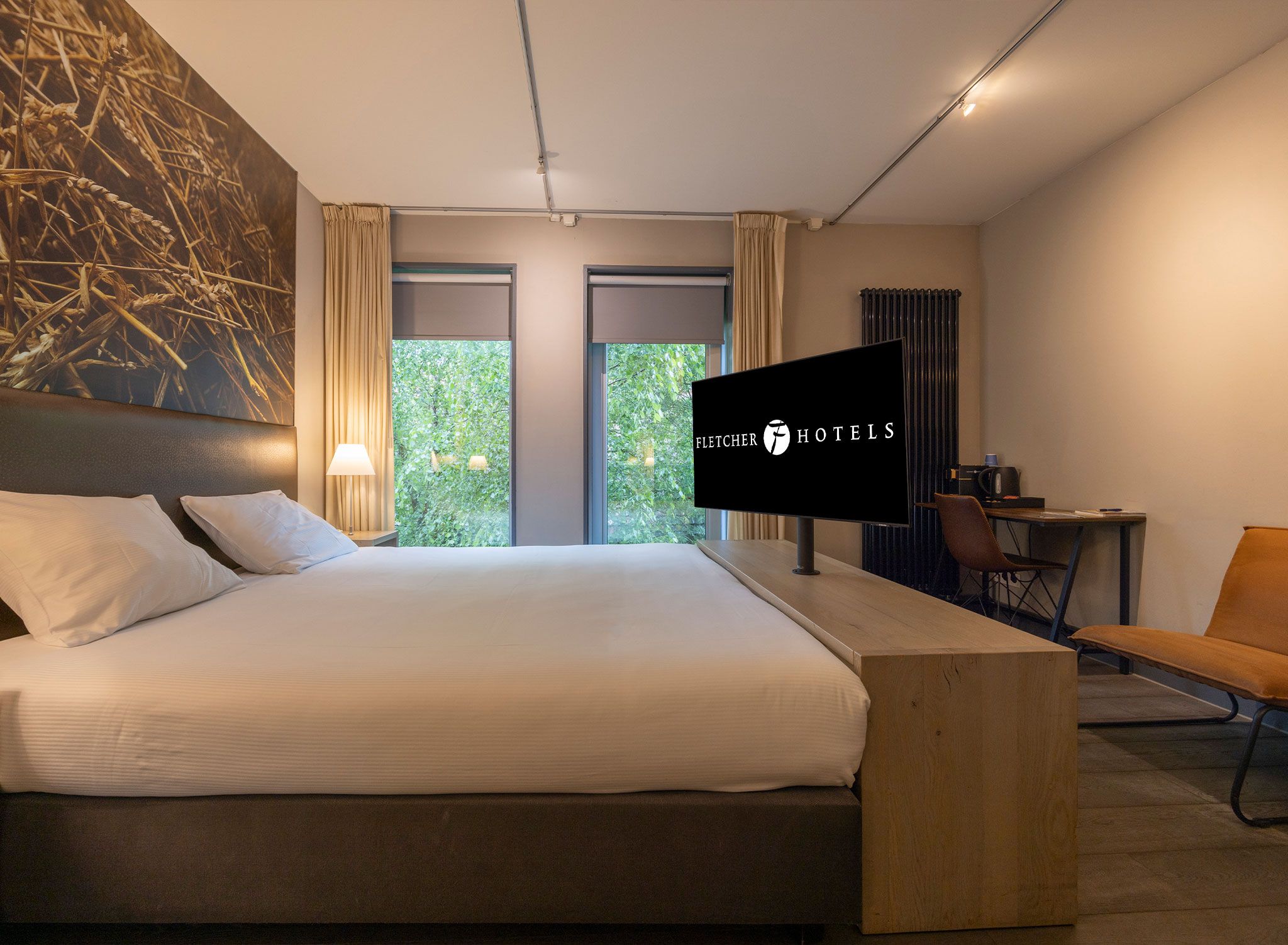 Ein Hotelzimmer mit einem großen Bett, einem Schreibtisch, einem Sessel und einem Fernseher, der das Logo von Fletcher Hotels zeigt.