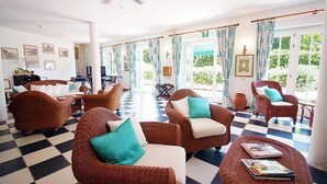 Un vestíbulo de hotel con mobiliario de mimbre, suelo a cuadros y grandes ventanales con cortinas.