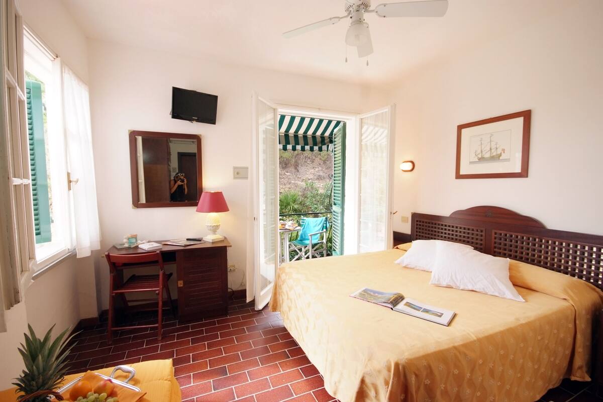A bedroom with a bed, a desk, a chair, a window, and a door leading to an outdoor area.