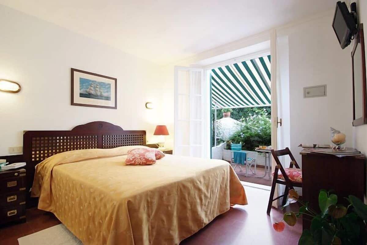 A bedroom with a bed, bedside table, lamp, and a view of the outside.