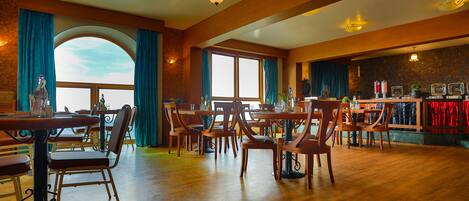 Un comedor espacioso con mesas y sillas de madera, ventanas amplias con cortinas y una pared de piedra al fondo.