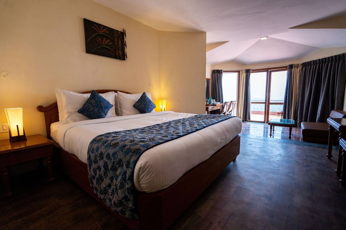 A spacious bedroom with a large bed, a wooden nightstand, a desk, and a balcony with curtains.
