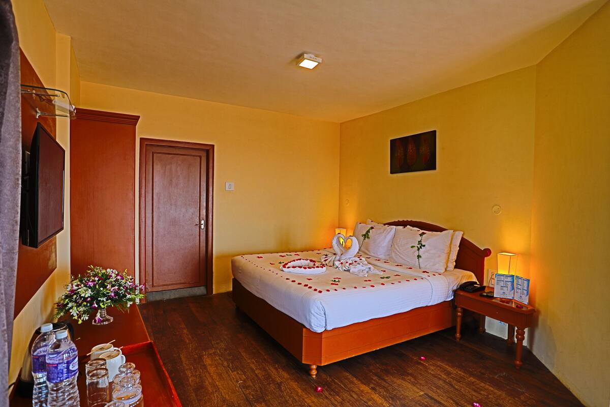 A hotel room with a wooden bed, a bedside table with a lamp, a small table with water bottles and glasses, and a door.