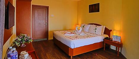 A hotel room with a wooden bed, a bedside table with a lamp, a small table with water bottles and glasses, and a door.
