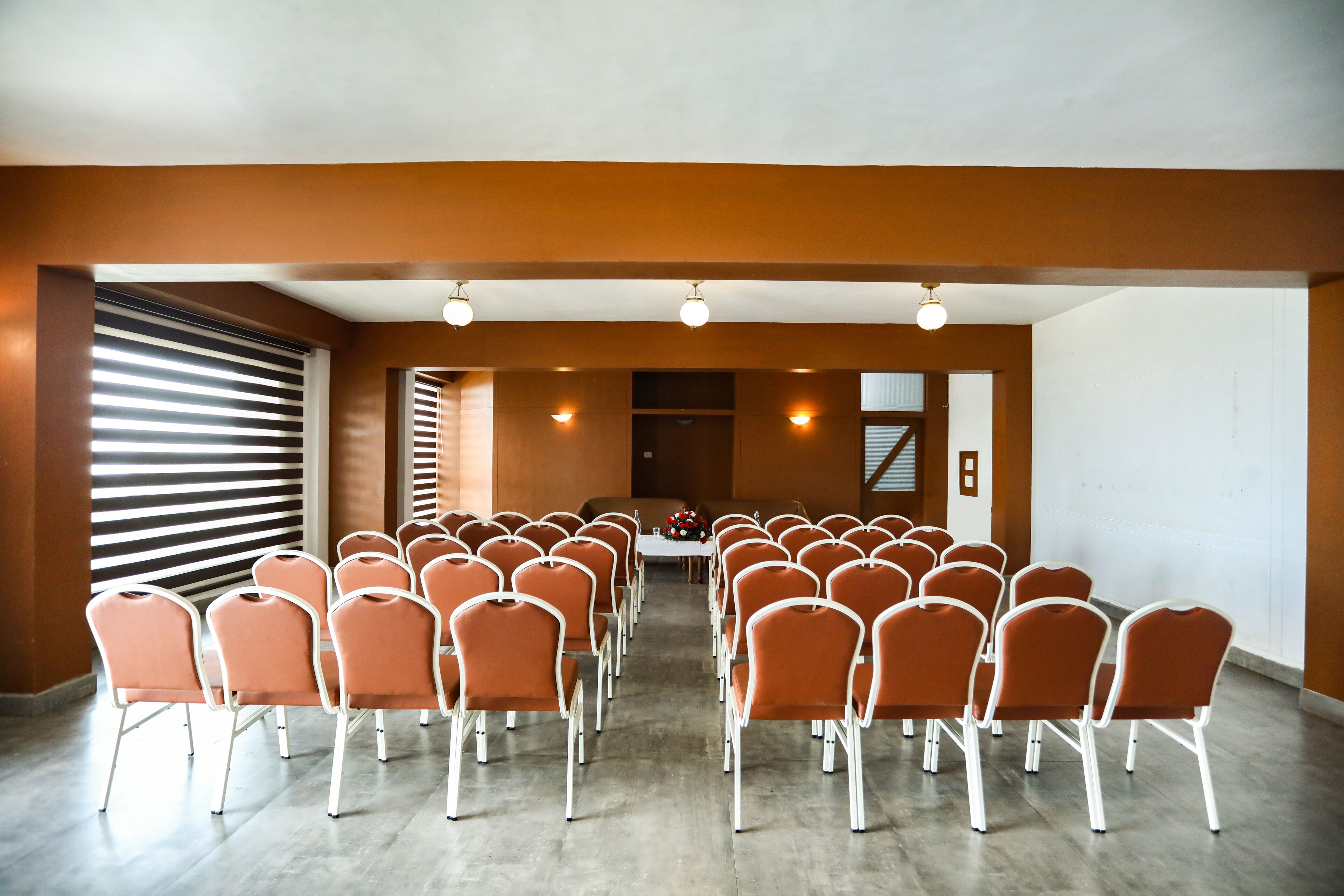 Une salle de conférence avec des rangées de chaises orange, une table blanche agrémentée d’un arrangement floral et une grande fenêtre dotée de stores.