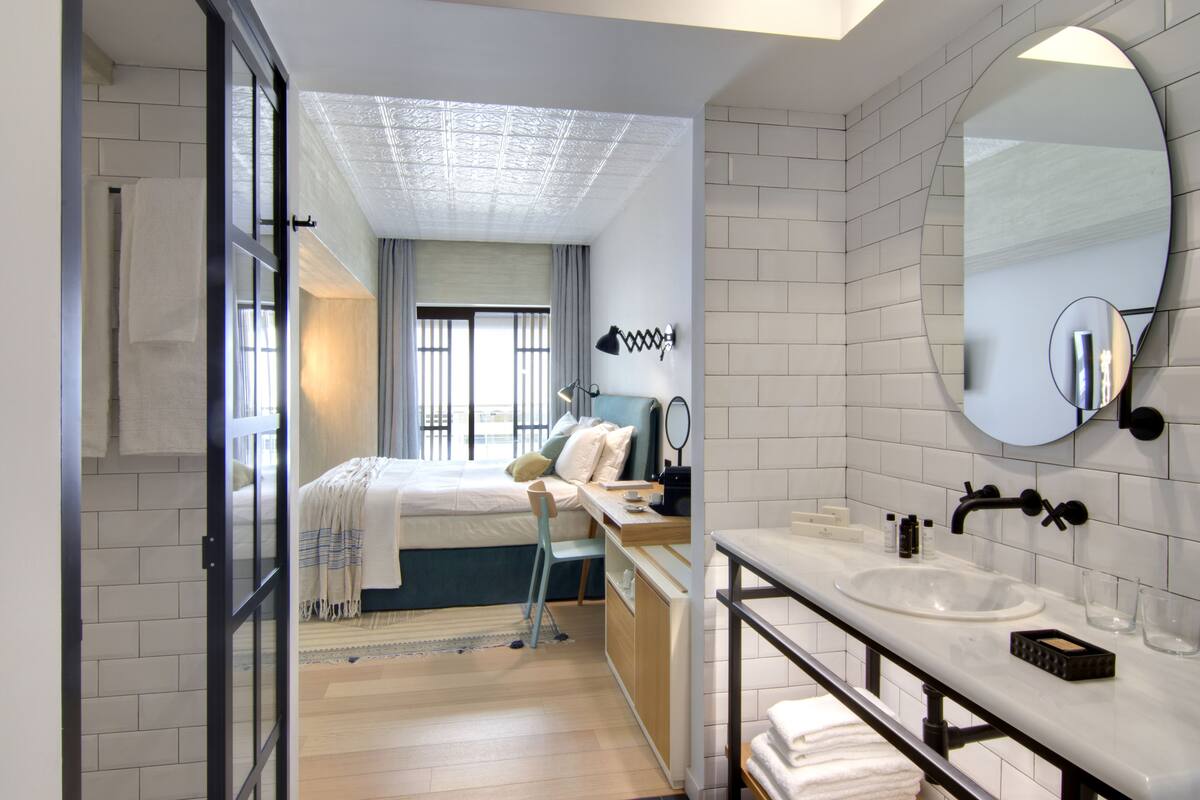 Quarto de hotel moderno com uma cama grande, uma escrivaninha, um banheiro com pia e espelho, e vista para a cidade pela janela.