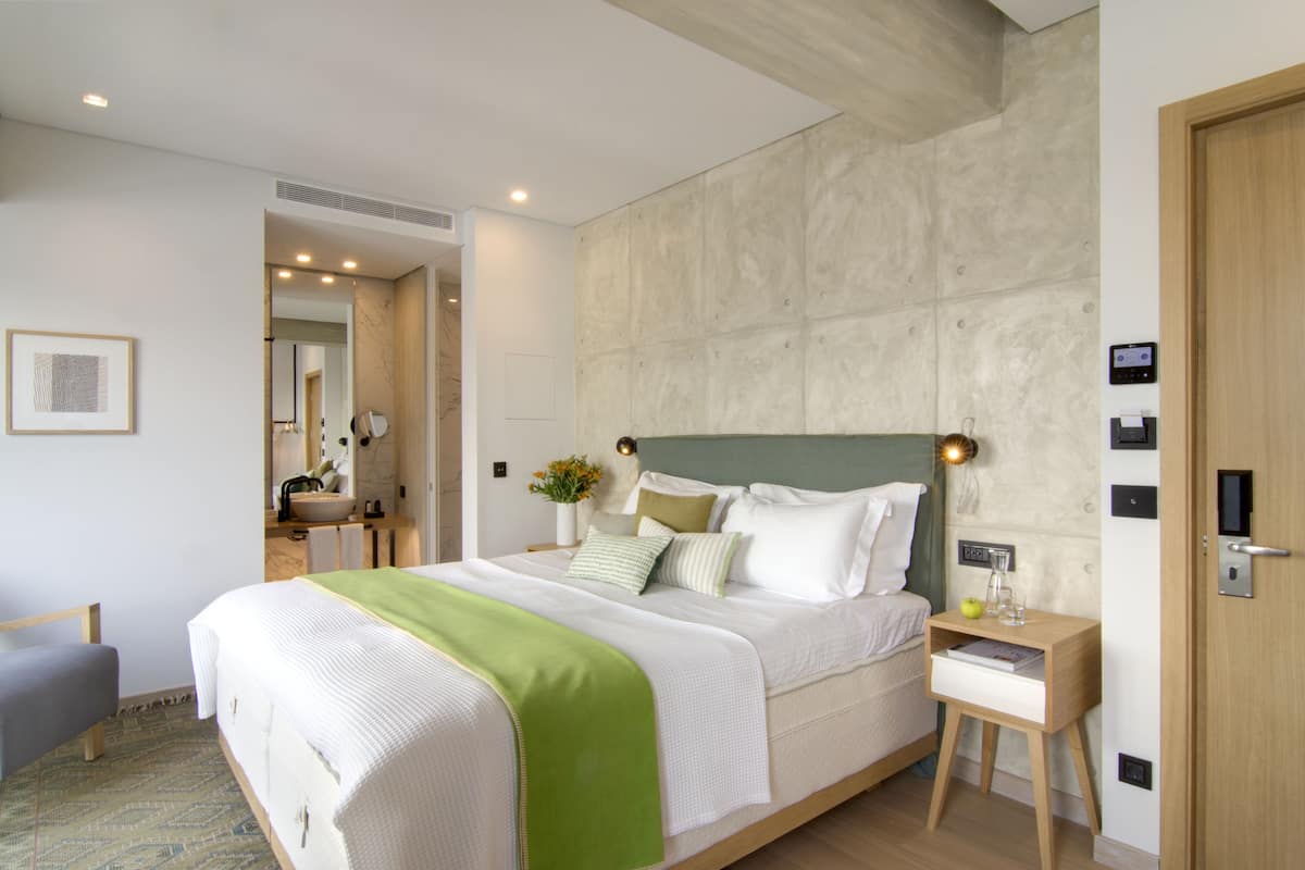 Quarto de hotel moderno com uma cama grande, mesas de cabeceira, uma mesinha de madeira e um banheiro visível através de uma porta aberta.
