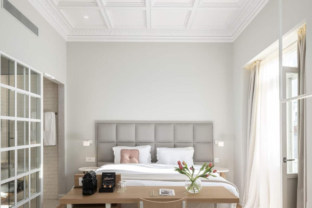 Quarto de hotel moderno com uma cama grande, uma escrivaninha e uma cadeira. Há uma janela com cortinas, um tapete estampado e um teto decorativo.