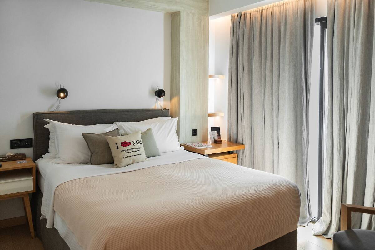 Uma cama bem arrumada com edredom bege e almofadas, uma mesa de cabeceira com abajur e uma janela com cortinas.