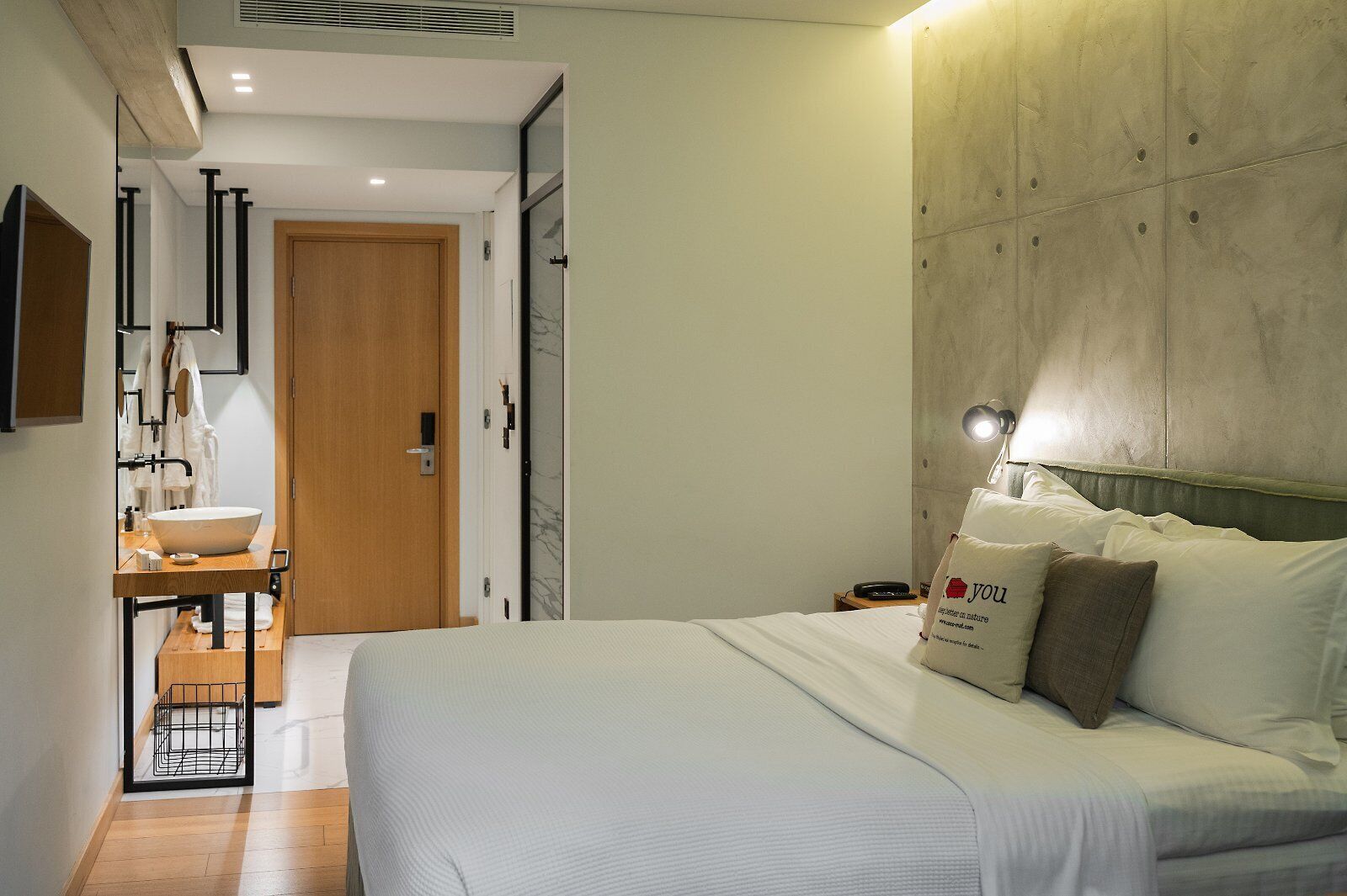 Quarto de hotel moderno com uma cama grande, uma mesinha de cabeceira, um espelho e cabeceira integrada.