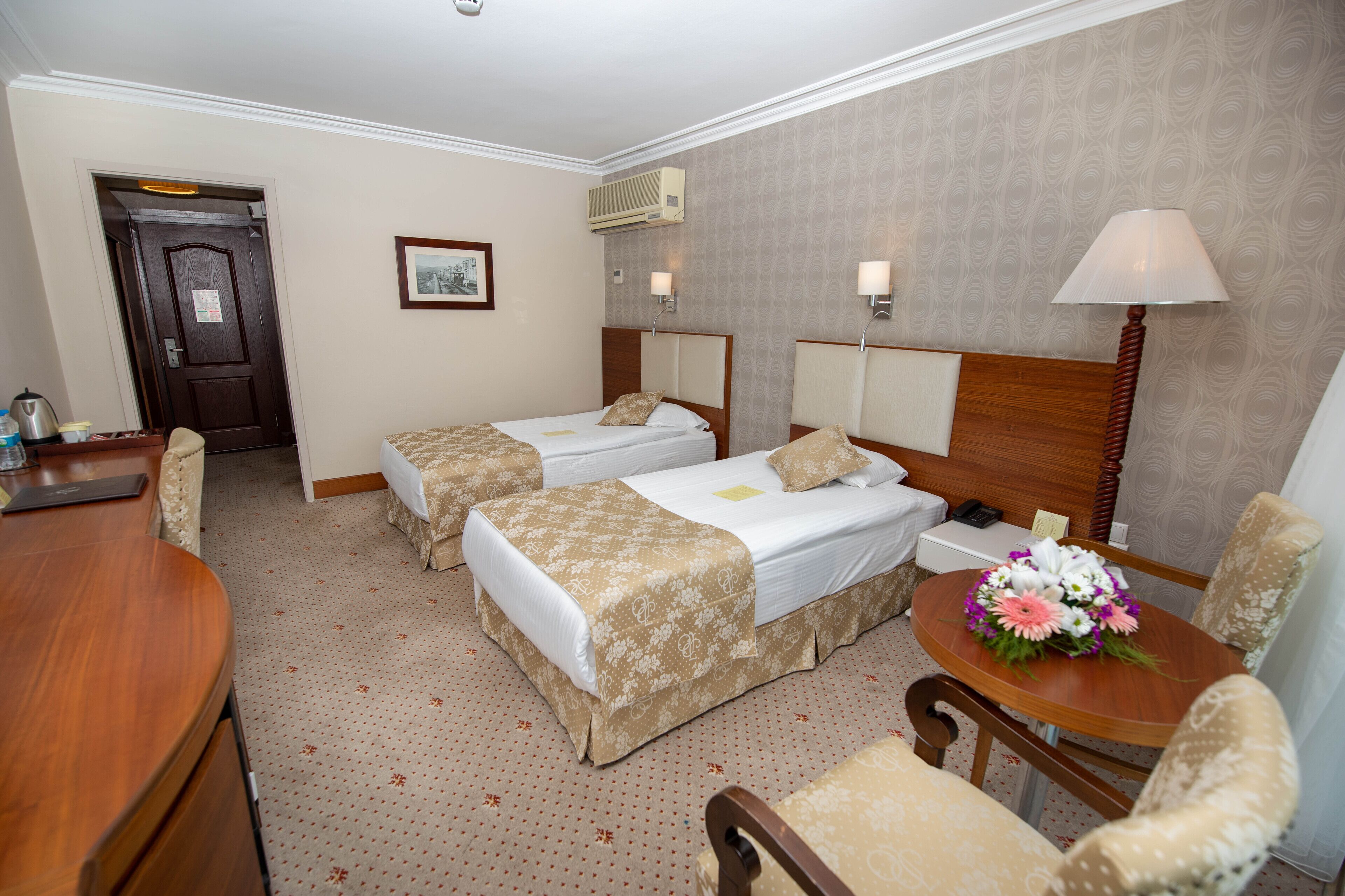 Habitación de hotel con dos camas individuales, un escritorio, una silla, una mesa con un arreglo floral y una puerta.