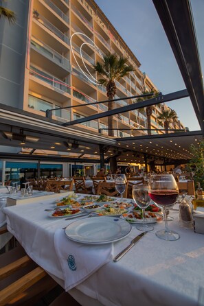 An outdoor dining area with tables set for a meal, palm trees, and a building with illuminated lines in the background. - Izmir Palas Otel (Izmir)