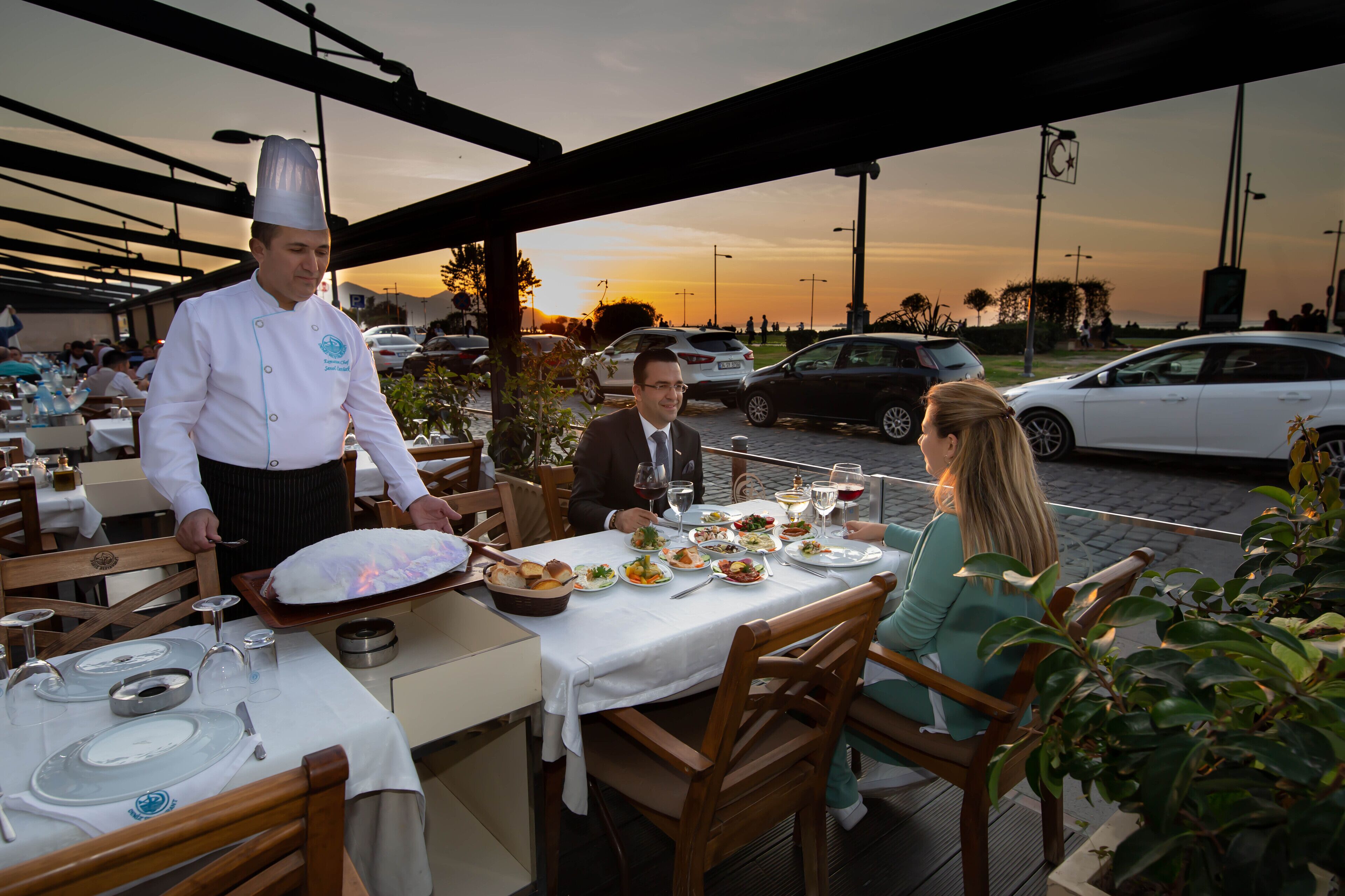 A restaurant with outdoor seating, a waiter serving food, and a sunset in the background.