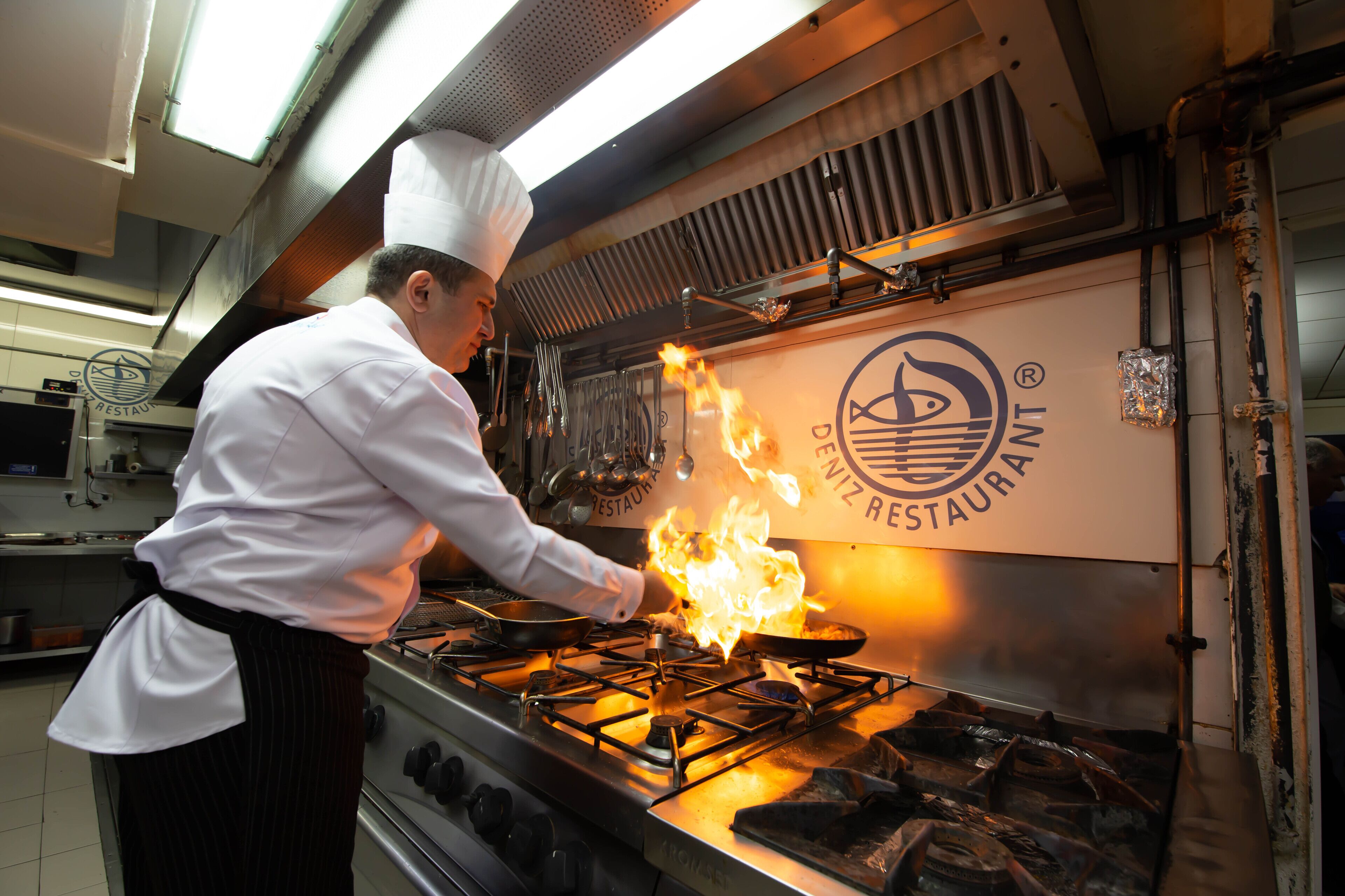 A chef performing a flambé with a pan on a stove in a commercial kitchen.