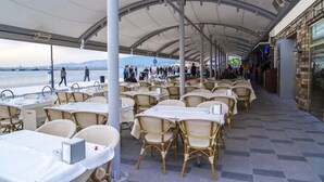 An outdoor dining area with tables and chairs, a covered walkway, and a view of the water and distant mountains. - Kilim Otel Izmir (Izmir)