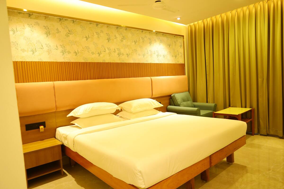 A hotel room with a large bed, a nightstand, a green armchair, a small table, and a curtain.