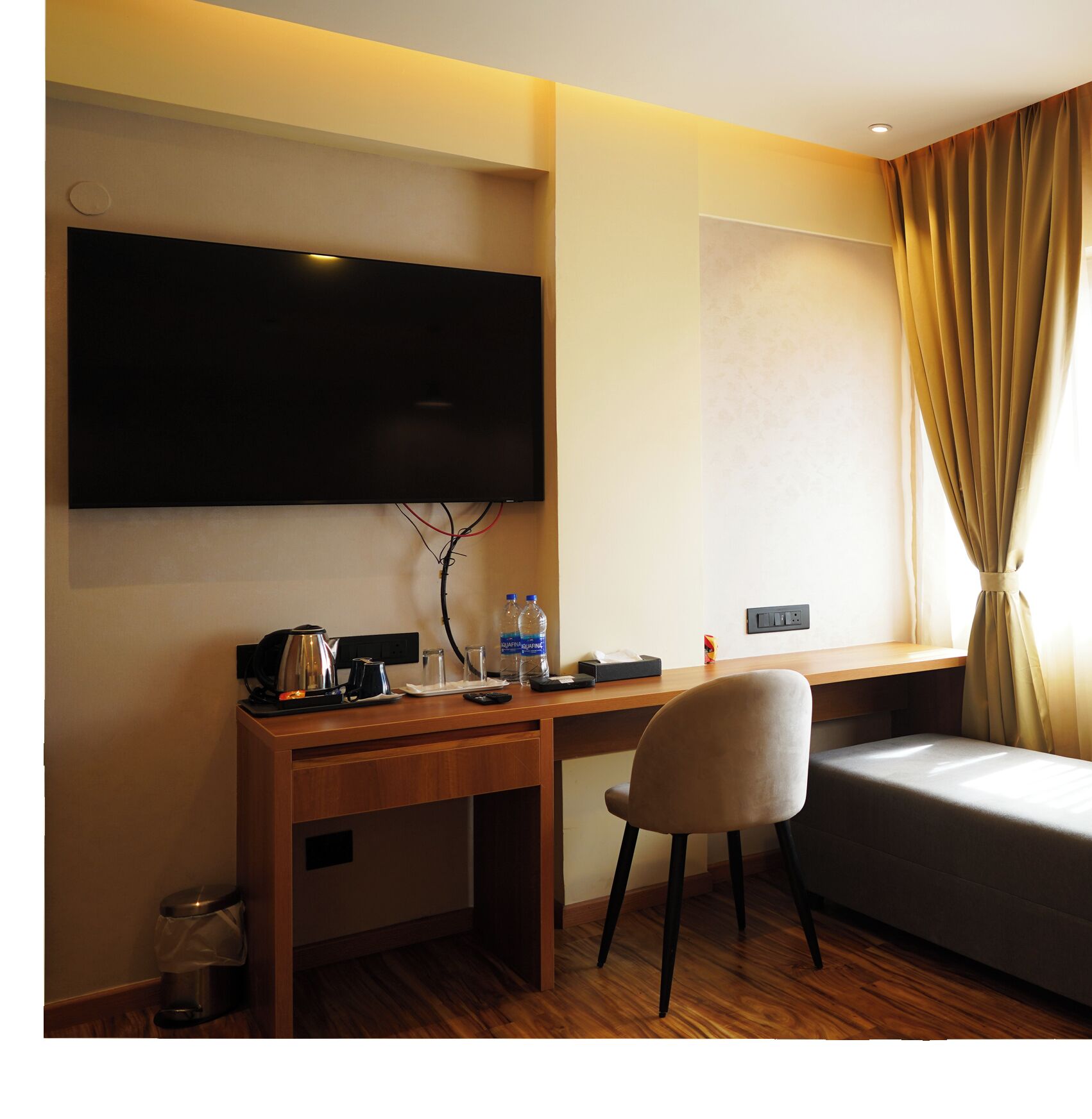 A hotel room with a wooden desk, a TV mounted on the wall, a bed, a chair, and a window with curtains.
