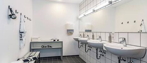 A modern bathroom with three sinks, a bench with towels, and a mirror with lights.