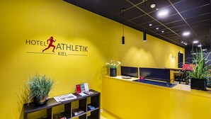Un mostrador de recepción con una pared amarilla, el logotipo del Hotel Athletik Kiel y una planta a la izquierda.