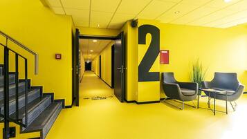 Un pasillo amarillo brillante con el número '2' grande en la pared, una escalera y dos sillas negras con una pequeña mesa.