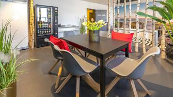 A modern dining area with a dark wooden table, grey chairs, and a vase of yellow flowers. A refrigerator and a vending machine are visible in the background.